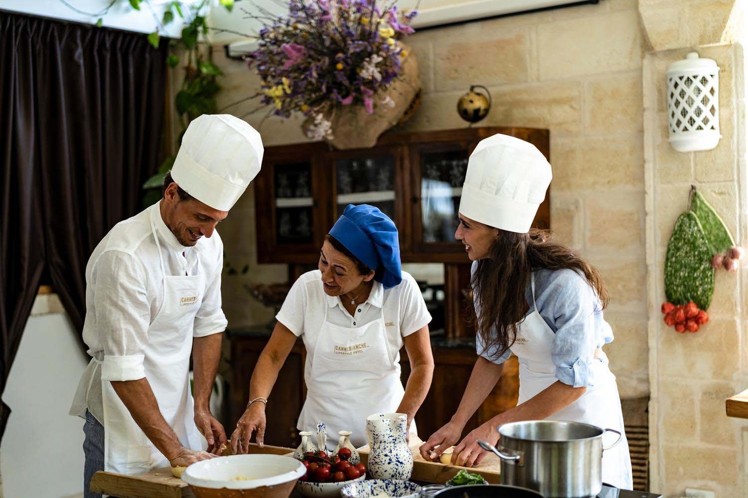A Apulian woman in a blue chef's hat stands between a man and woman, wearing white chefs' hats, with bowls, pans and various ingredients on the table. She is showing them how to cook a local dish.
