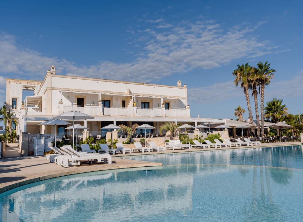 A white villa-style building overlooks sun beds around a large, curved pool against a background of palm trees and blue skies.