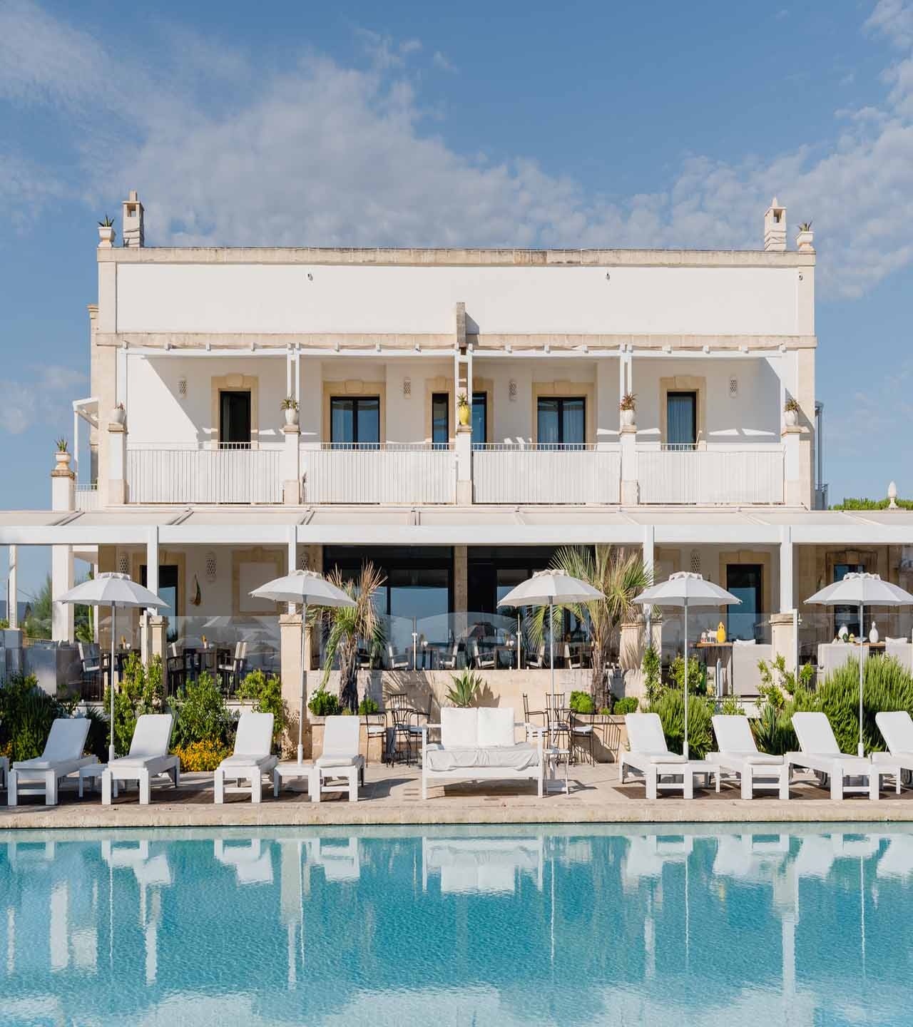 A whitewashed hotel building overlooks sun beds lined up by a large pool against a background of palm trees and blue skies.