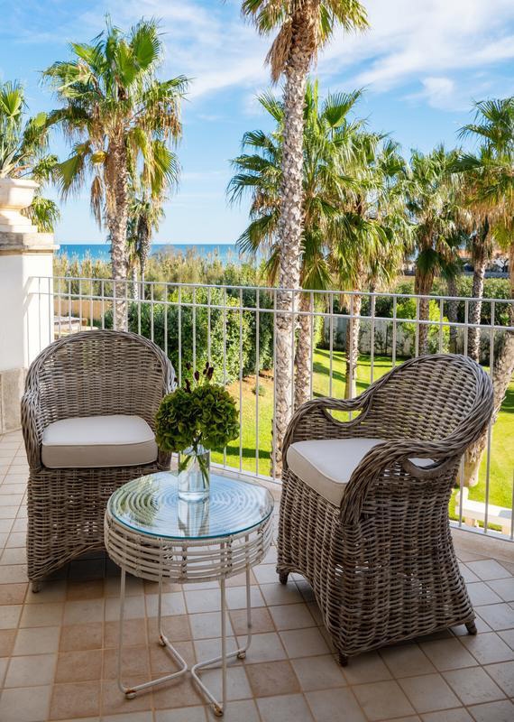 Two rattan chairs and a glass topped table on a terracotta tiled terrace, with railings and views of palm trees and gardens with the sea in the near distance.