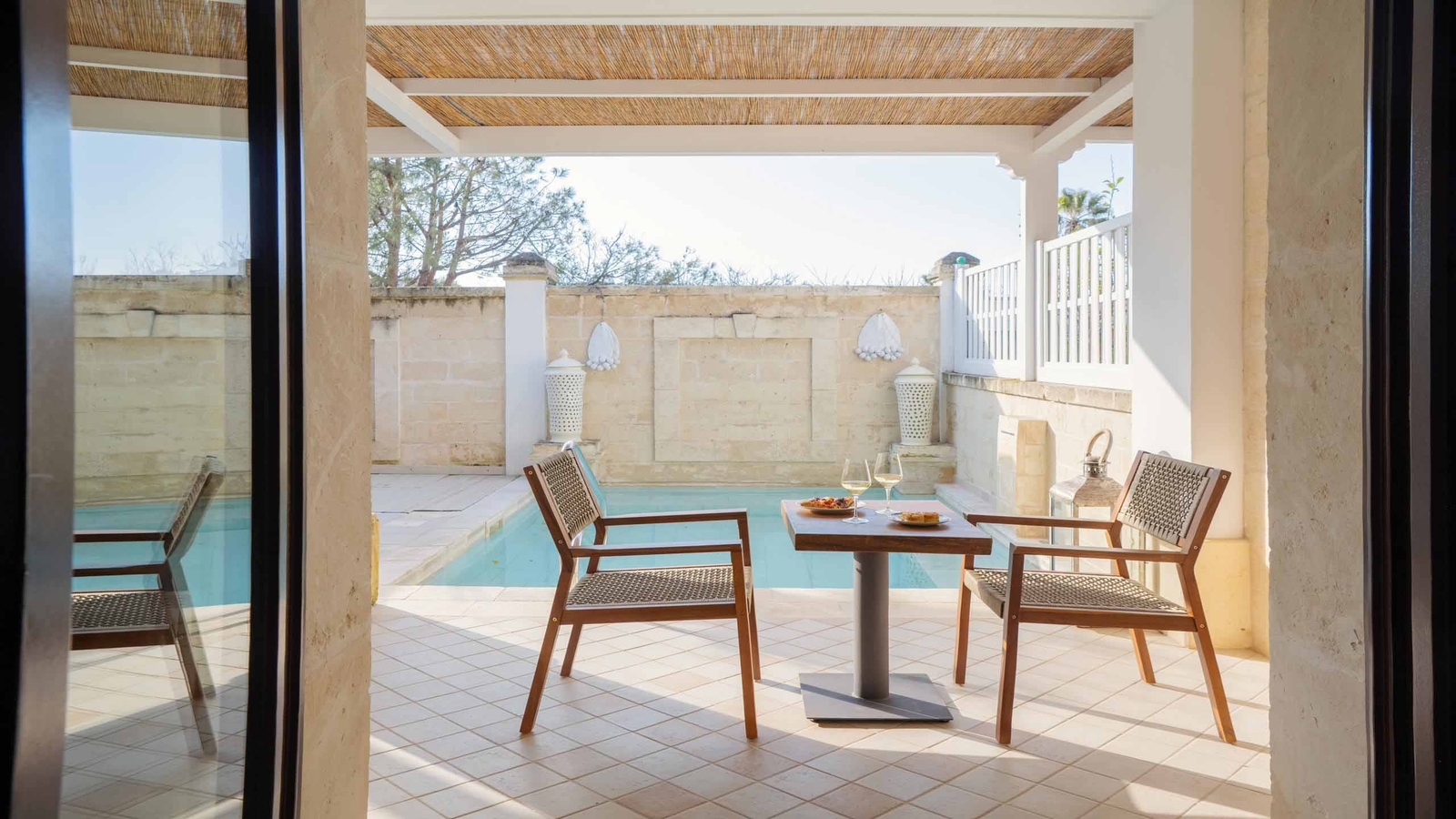 A wooden table and two chairs with plates of food and glasses of white wine, on a terrace by a small private pool, viewed from inside a room with sliding glass doors.