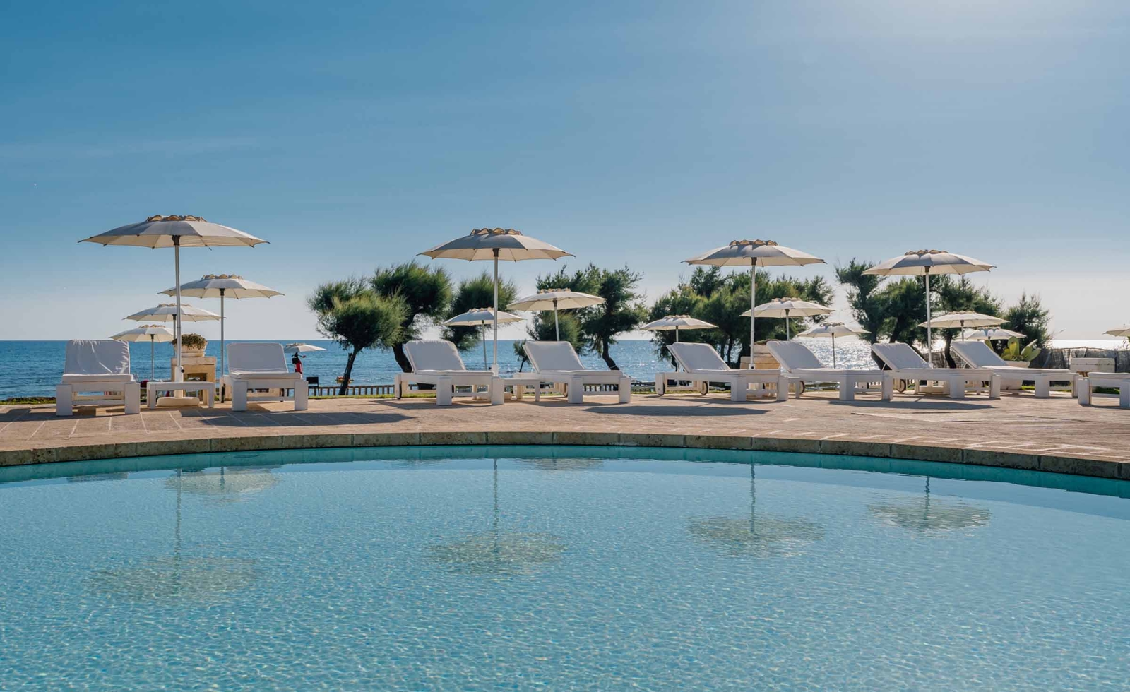 A curved pool in the sunshine with sun beds and umbrellas behind, blue skies and views of the sea.