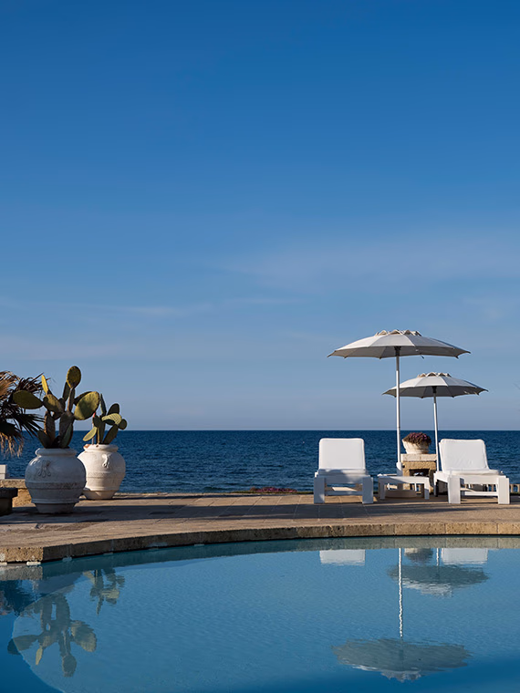 The curved edge of a swimming pool under blue skies with two sun beds, umbrellas, potted plants and sea views.