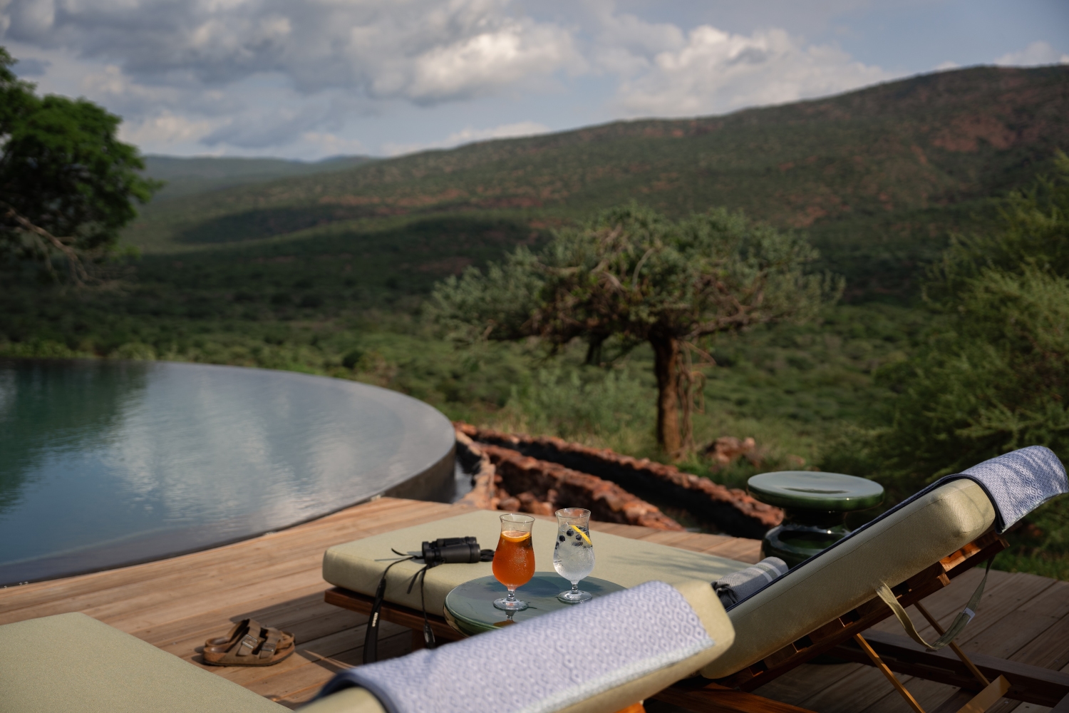Exterior view of two sun loungers facing a round infinity pool overlooking a baobab tree in the foreground and a green valley and mountains beyond.