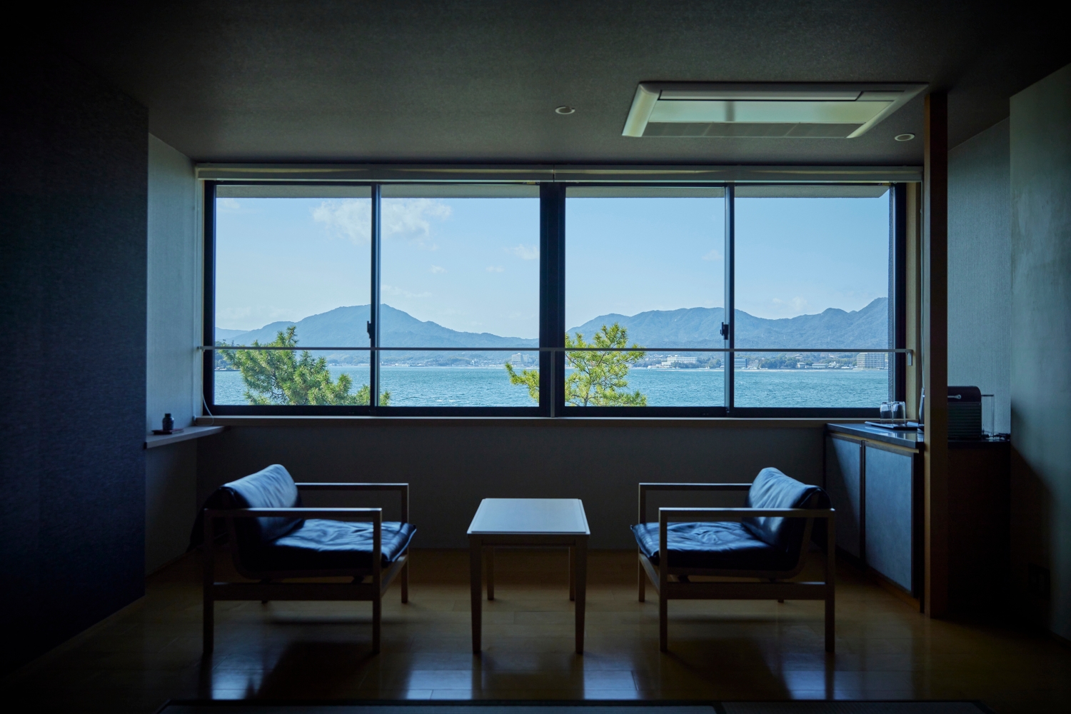 Interior view of a seating area with two vintage chairs facing a small coffee table in front of a long picture window overlooking the sea and mountains.