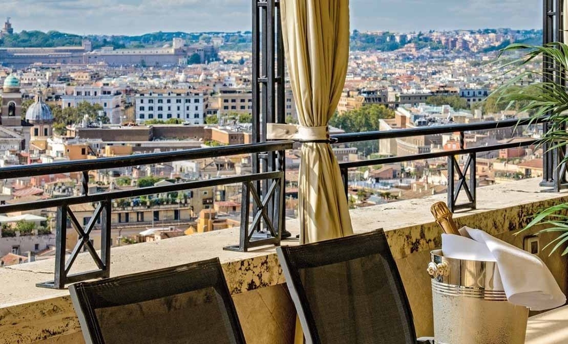 An open-sided balcony has two chairs and a bottle of champagne on a table, facing views of Rome's rooftops.