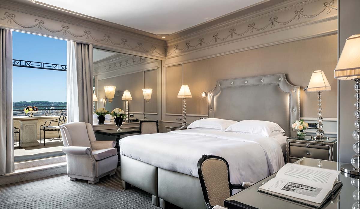 A hotel bedroom decorated in white and off-white tones, with a double bed and armchair and an open door to the balcony and views of Rome.