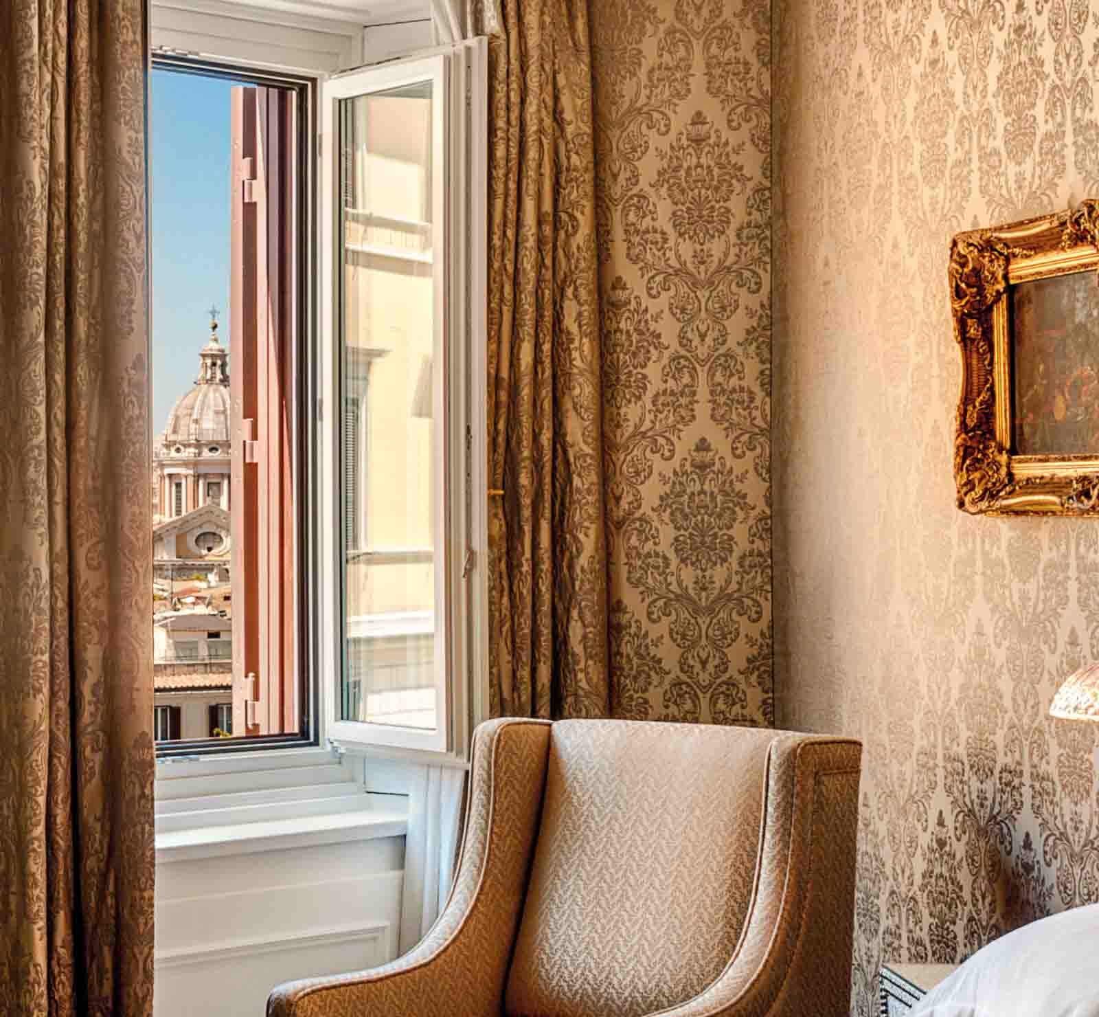 A taupe chair, gold flocked wallpaper and matching curtains by an open window with views of Rome's rooftops.