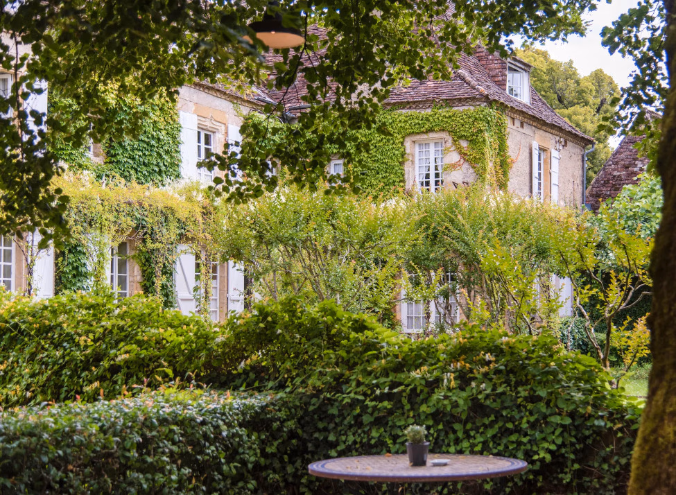 Exterior view of a stone manor house covered in ivy with green hedges and a small table in the foreground.