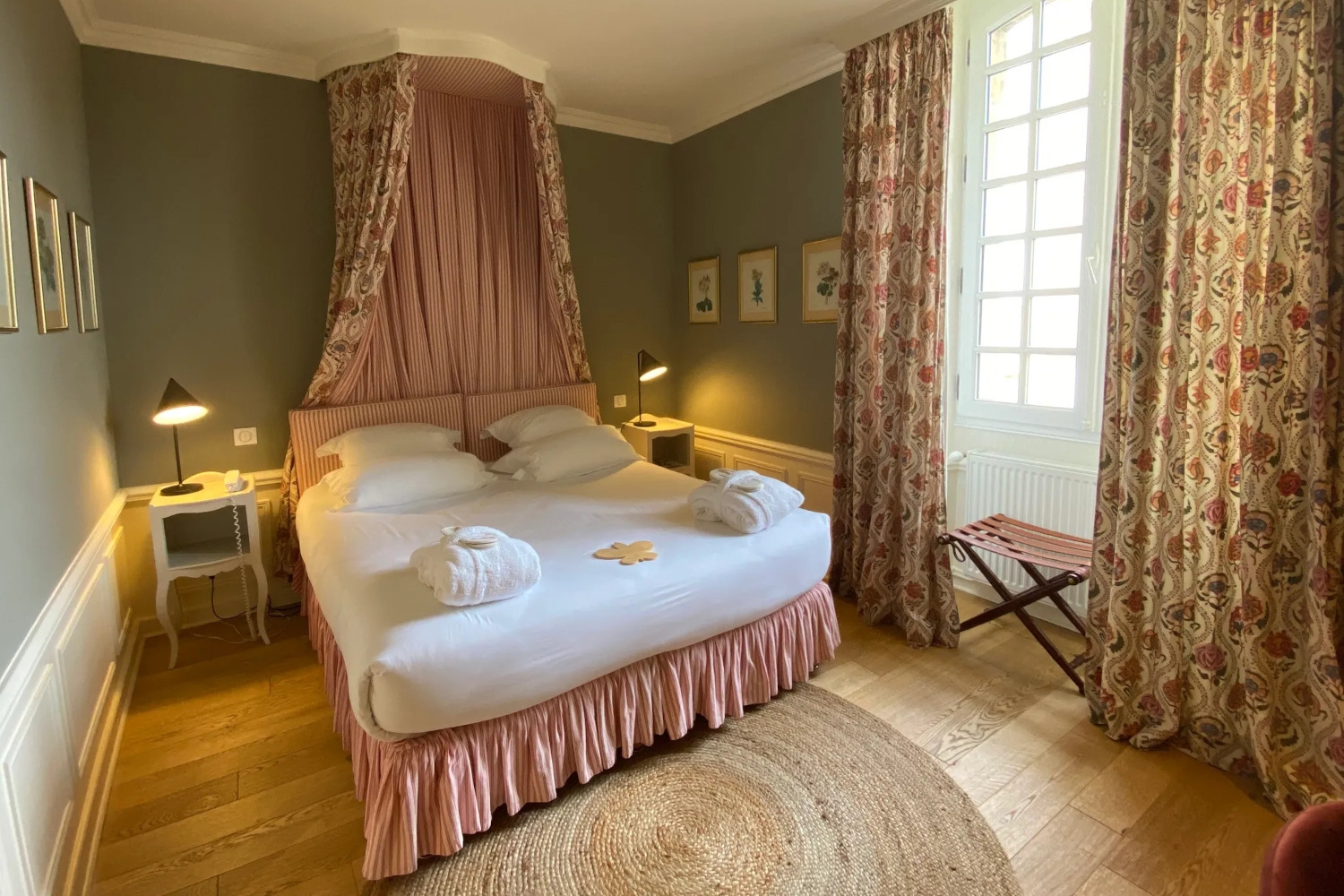 View of a bedroom with a bed with red canopy and white sheets, and a window with long curtains to the right, and hardwood floors, and bedside tables with lamps on.