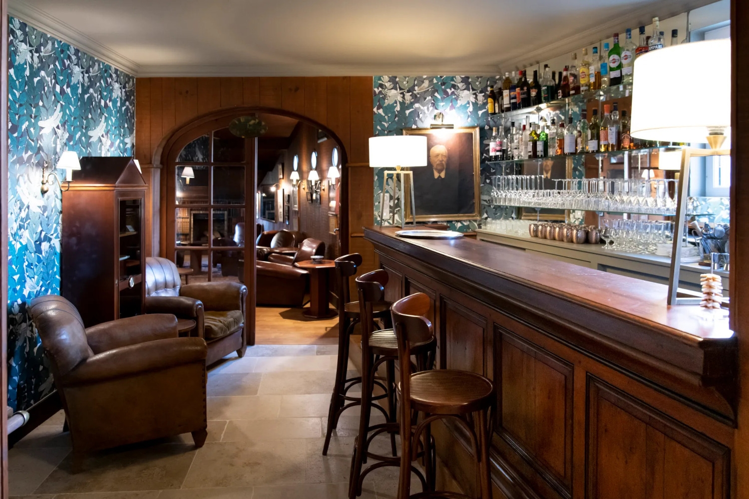 Interior of a dark wood bar with three high-top bar stools and a lamp lit on the bar, and lots of bottles on the wall behind.