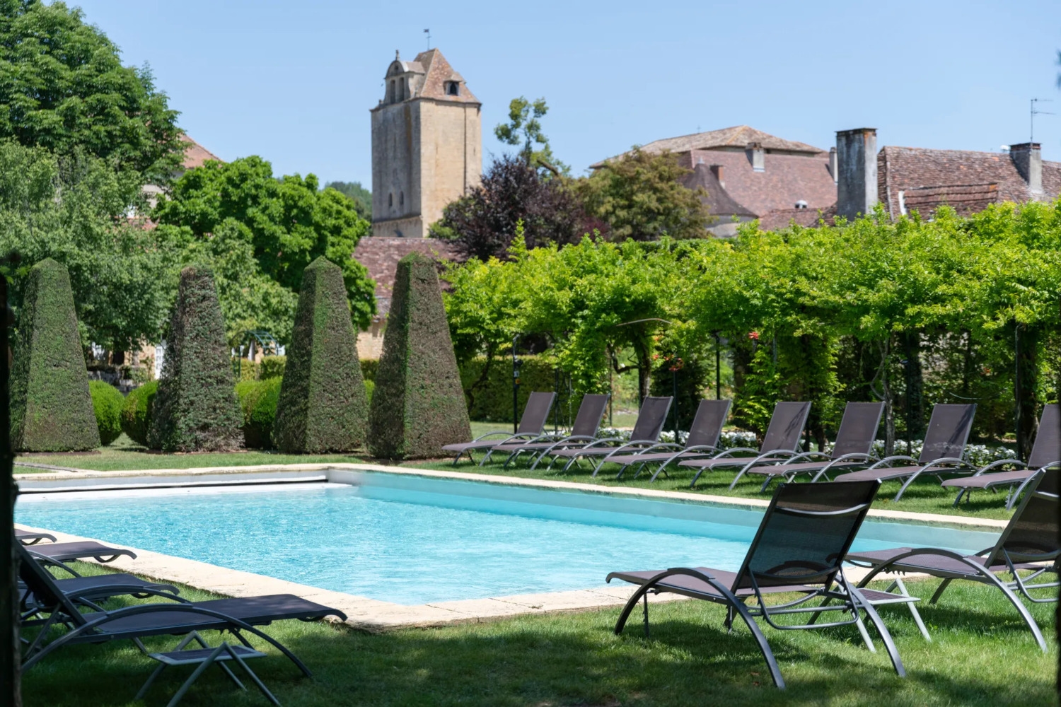 Exterior shot of a swimming pool with one lounge chair in the foreground and several lounge chairs beyond it as well as two pyramid shaped hedges and a stone manor with a tower rising in the background.