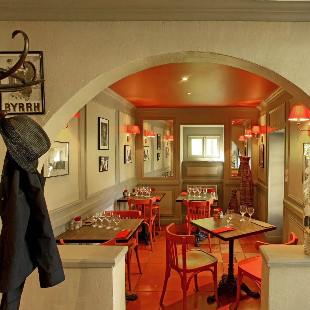 Interior view of a restaurant with a few tables and some red lamps casting red light on the floor, and in the foreground a hook with a man's hat and jacket.