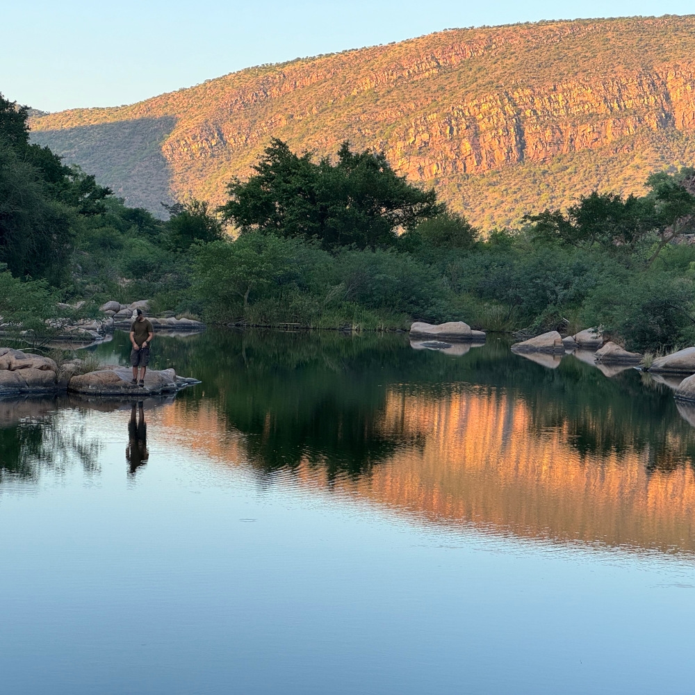 Man stands on a distant rock next to a large river or pool with trees in the near background and an illuminated mountain further distant.