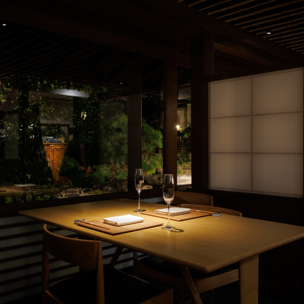Low-lit dining room with a simple wooden table in the foreground set for dinner, a paper screen in the background and a darkened garden outside a set of windows in the background.