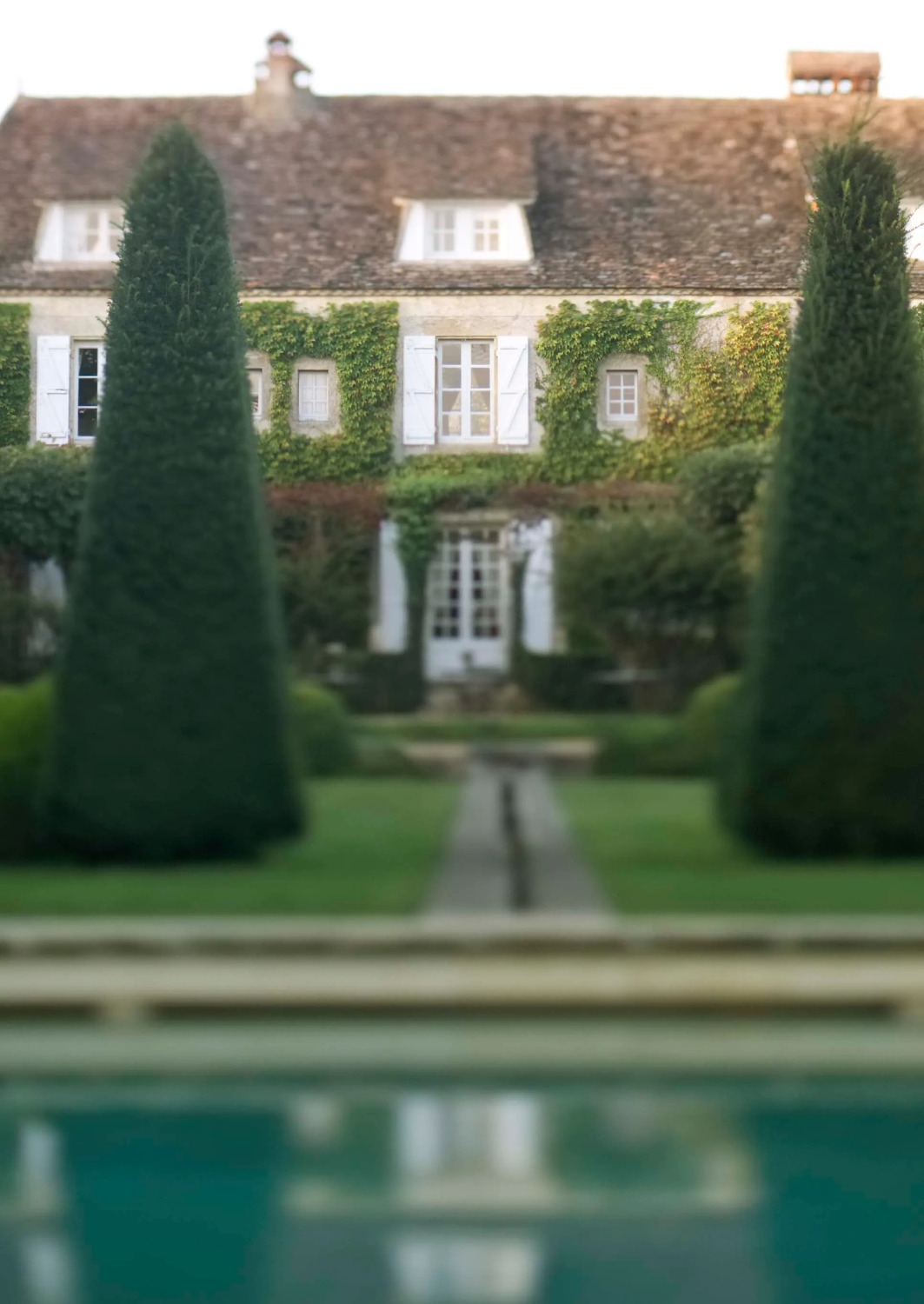 Exterior view of the front of a stone manor covered in ivy with a garden and pyramid-shaped hedges and lawn in the foreground.