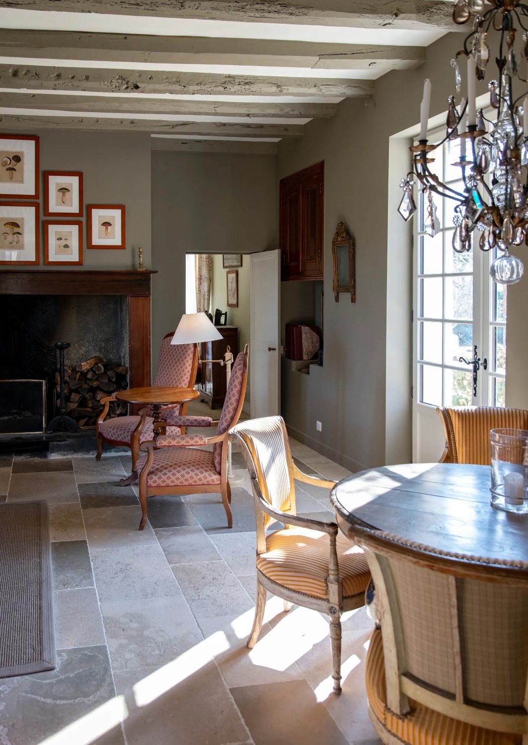 Interior view of a desk with two chairs in the foreground and two lounge chairs by a large fireplace in the background, with old ceiling beams and a large window on the right.