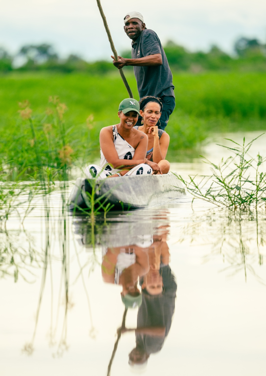 Two people in a small mokoro canoe being paddled by a safari guide at the back, floating through wetland waterway surrounded by green grass.