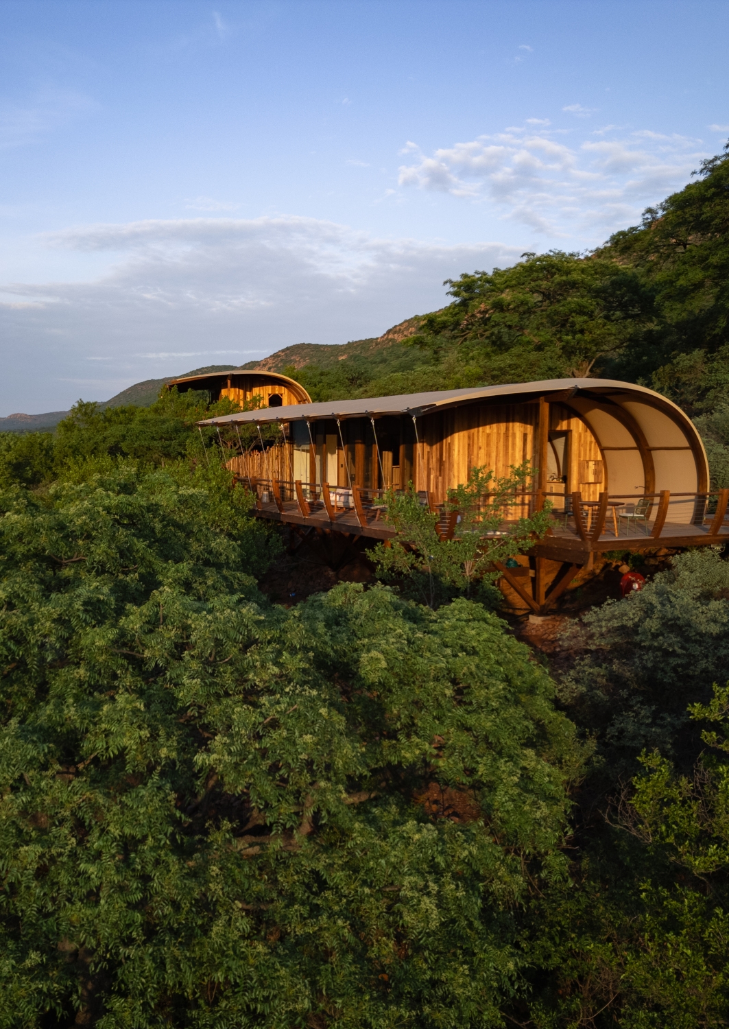 Exterior shot of two adjacent, cliffside safari suites with with canvas roof and wraparound wooden deck set among green trees with mountains in the distance.