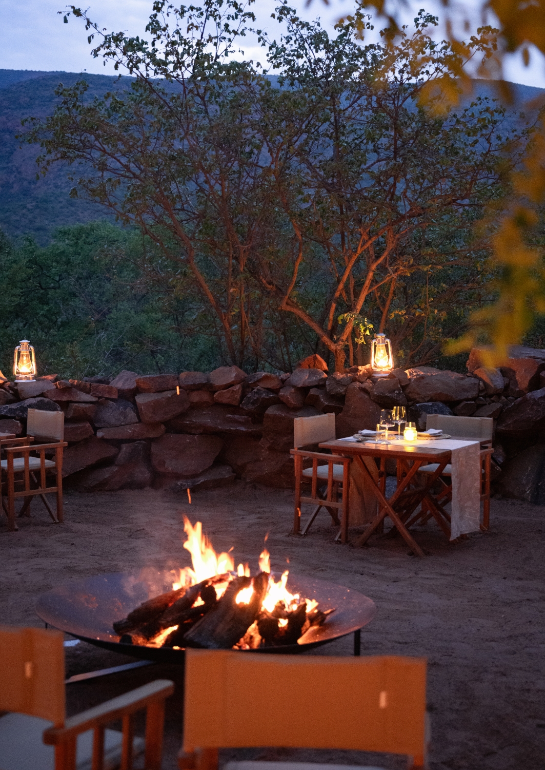 Exterior shot of a traditional boma yard encircled by a stone wall, with a small bonfire and a table set for dinner with a candle and lanterns, and low evening light.