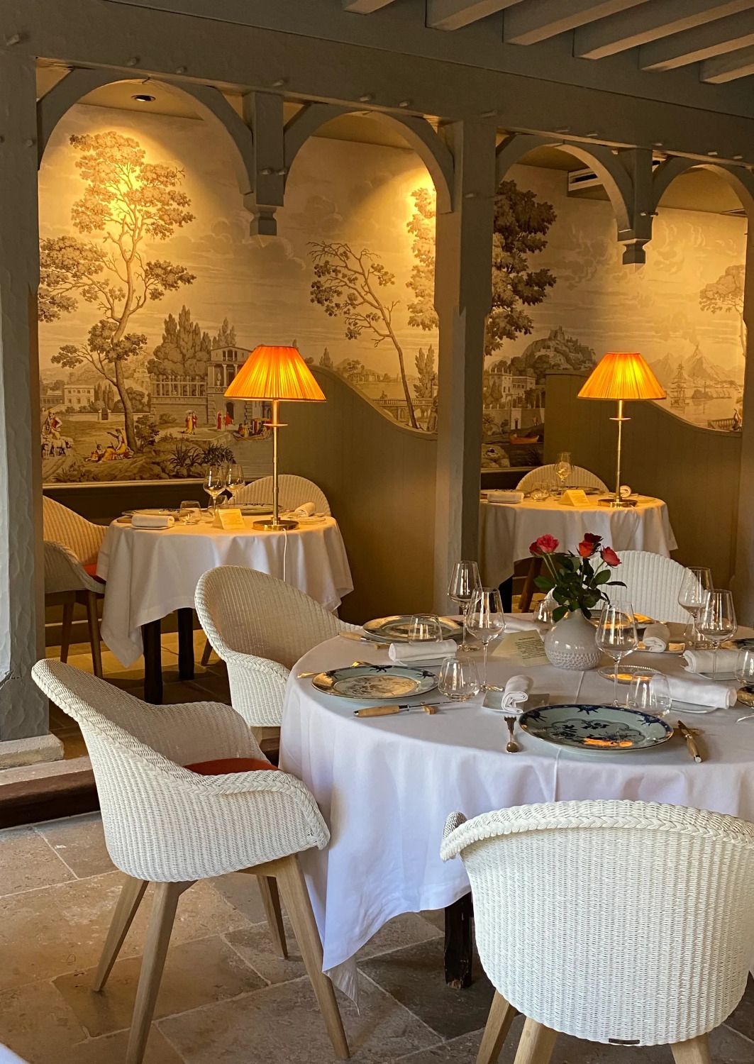 Interior shot of a formal restaurant dining area with white table cloth round tables and white chairs, and orange lamps, and wallpaper with trees in the background.