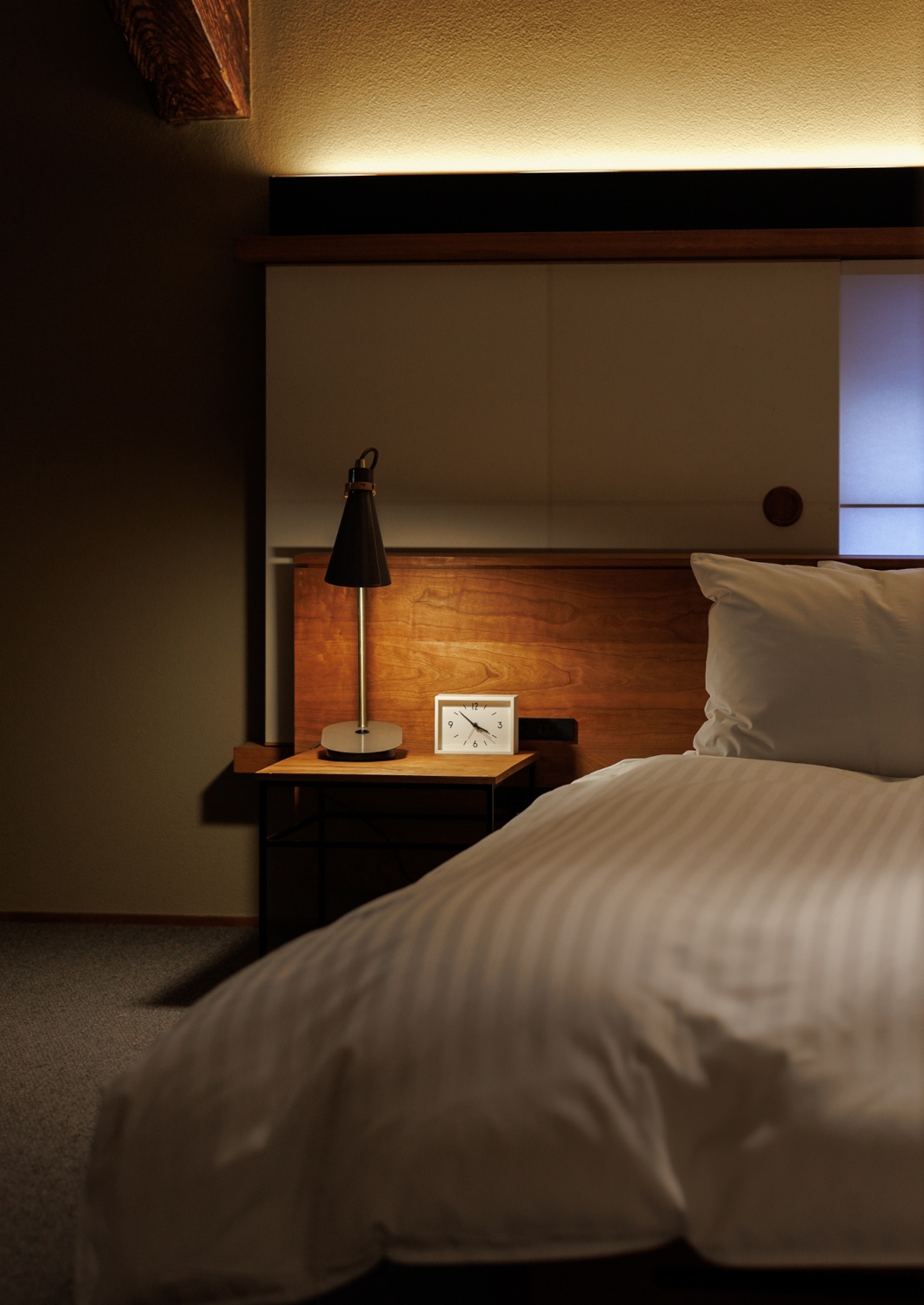 Interior of a ryokan berdroom with a western-style bed with white bedding and a small minimalist bedside table with a lamp on.