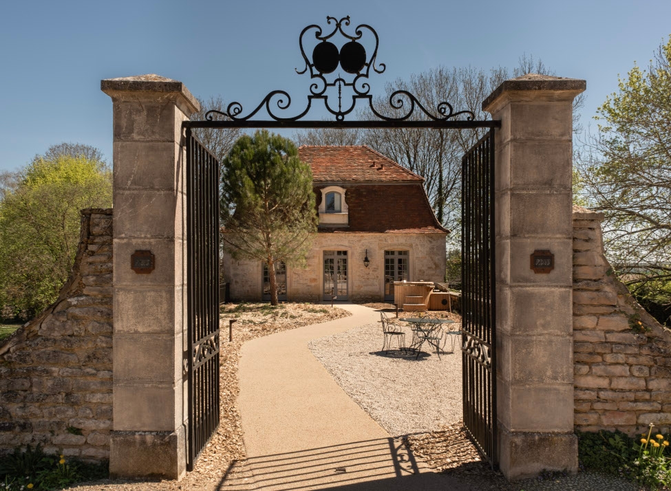 A stone gate with wrought-iron detailing above leads into a paved drive with a French country house at the end, surrounded by a few trees.