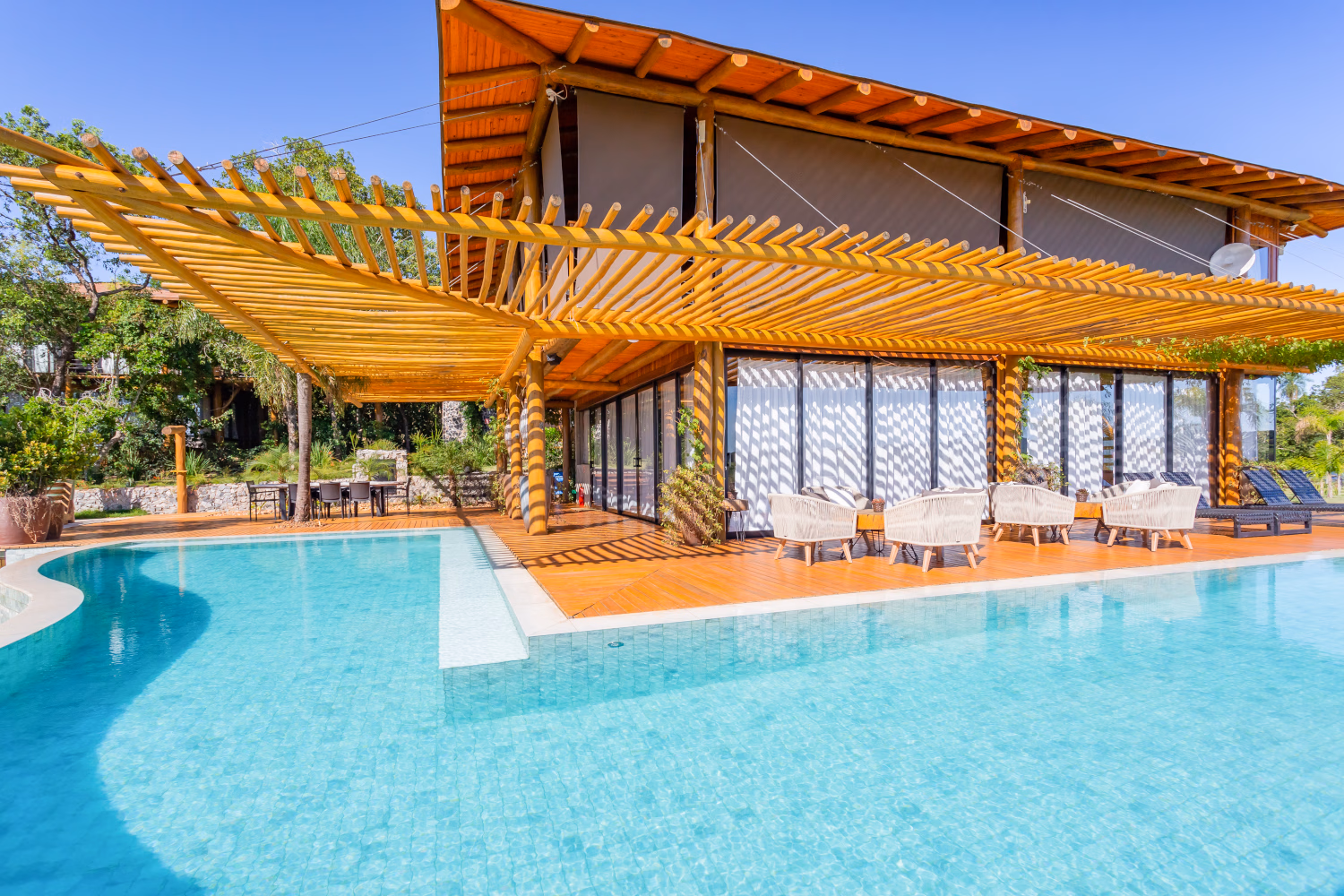 Exterior of the villa with an L-shaped wraparound pool in the foreground, wooden shading and the two-story villa and decking behind.