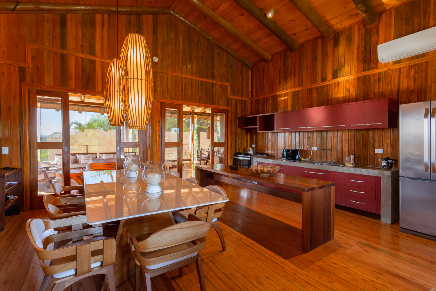 Interior of the Bonito Villa kitchen - a full wooden interior with dining table in the foreground, large hanging light fixtures and a galley kitchen area.