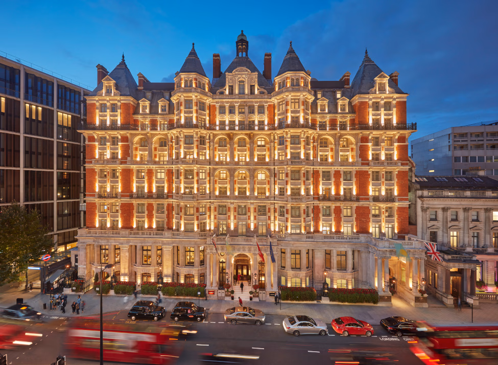 Exterior of a grand redbrick building with turrets in early evening with the front lights lit up, a dark evening sky and cars blurred in the foreground.