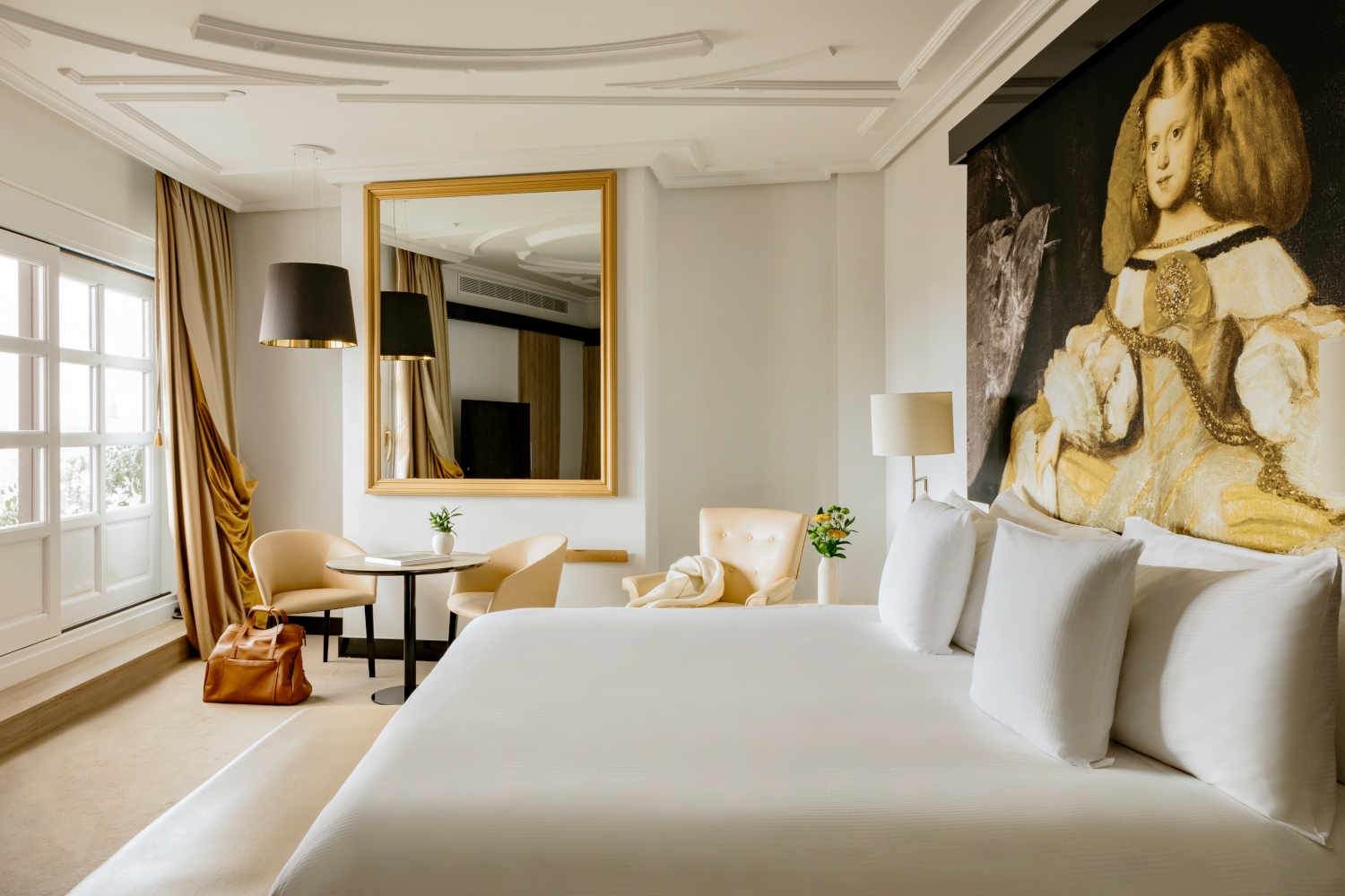 Interior shot of a bedroom with a white bed in the foreground and a large mirror and lamp with a black shade against the back wall.