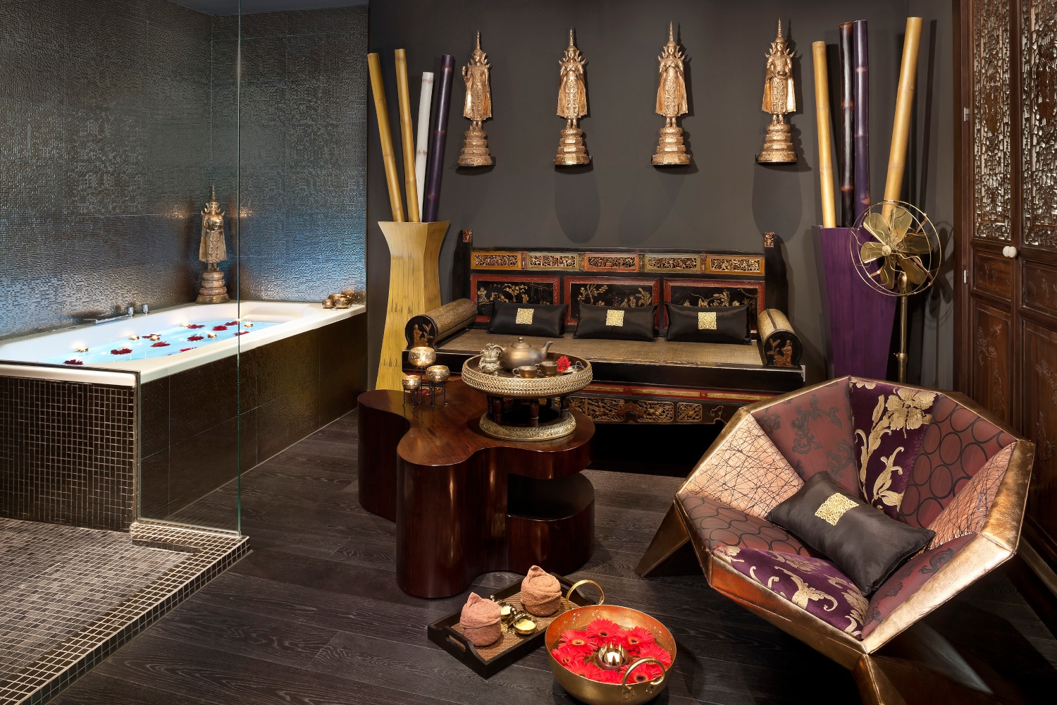 Interior of a spa with a tub, wooden carved sofa and relaxation areas decorated in dark colours with Thai decor.