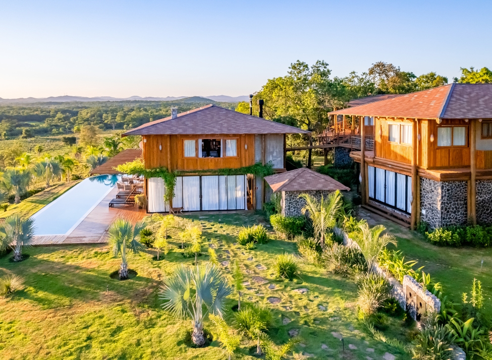 Exterior of two light brown villa buildings with an outdoor pool and surrounded by open green fields and a few trees.