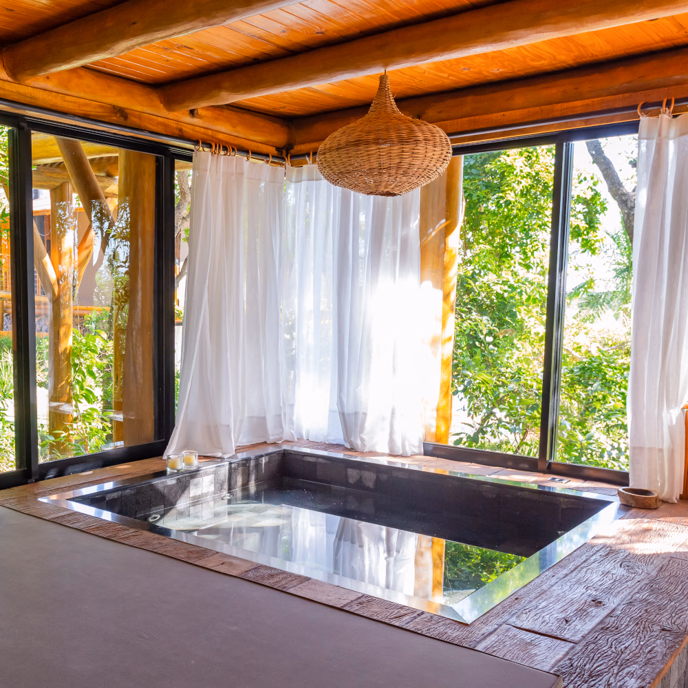 Interior of a small square plunge pool in a wooden room surrounded by floor-to-ceiling windows overlooking greenery.