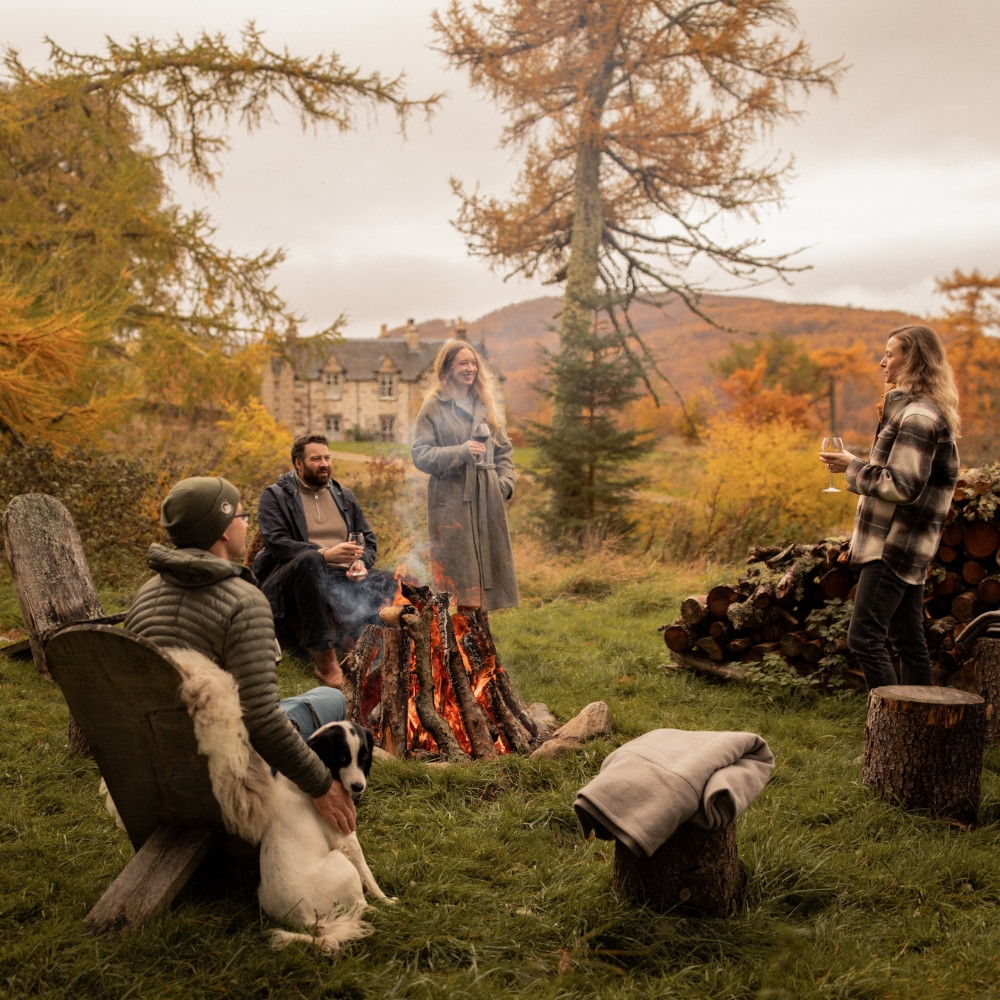 Four people sitting on carved wooden chairs around a fire pit in a small grassy clearing surrounded by autumn coloured trees.