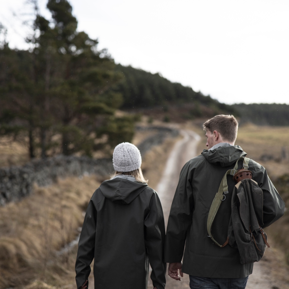 Two people dressed in hats and rain gear walking along a countryside track with a few trees in the distance.