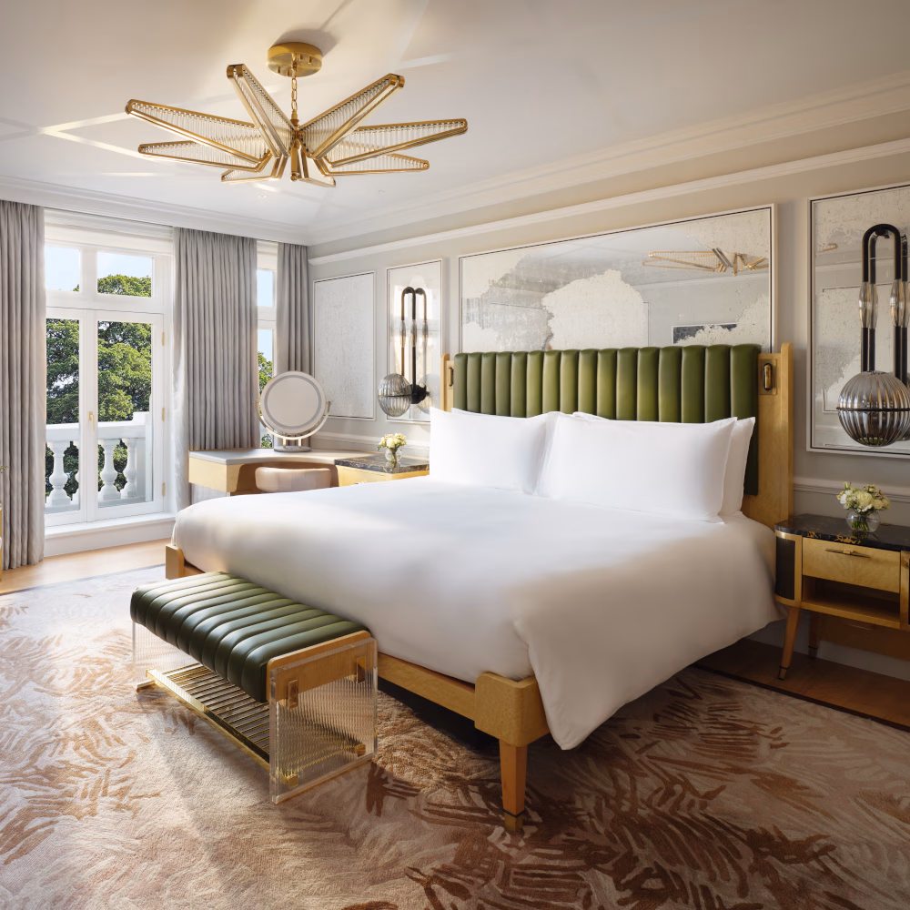 Interior of a bedroom with a white bed and green headboard and a window opening out onto a juliette balcony with white railing.