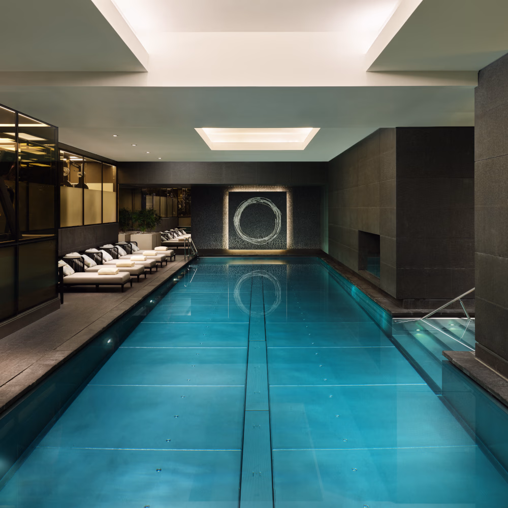 A long, rectangular indoor pool with dark grey walls and a line of white recliner chairs along the left side.