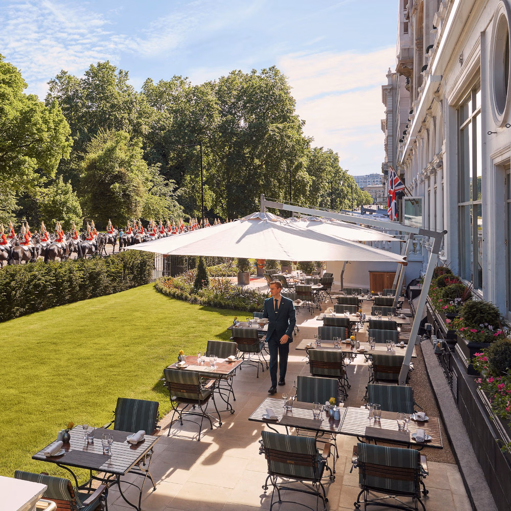 An outdoor patio seating area with tables and umbrellas overlooking a small lawn and, beyond, British guards on horses.
