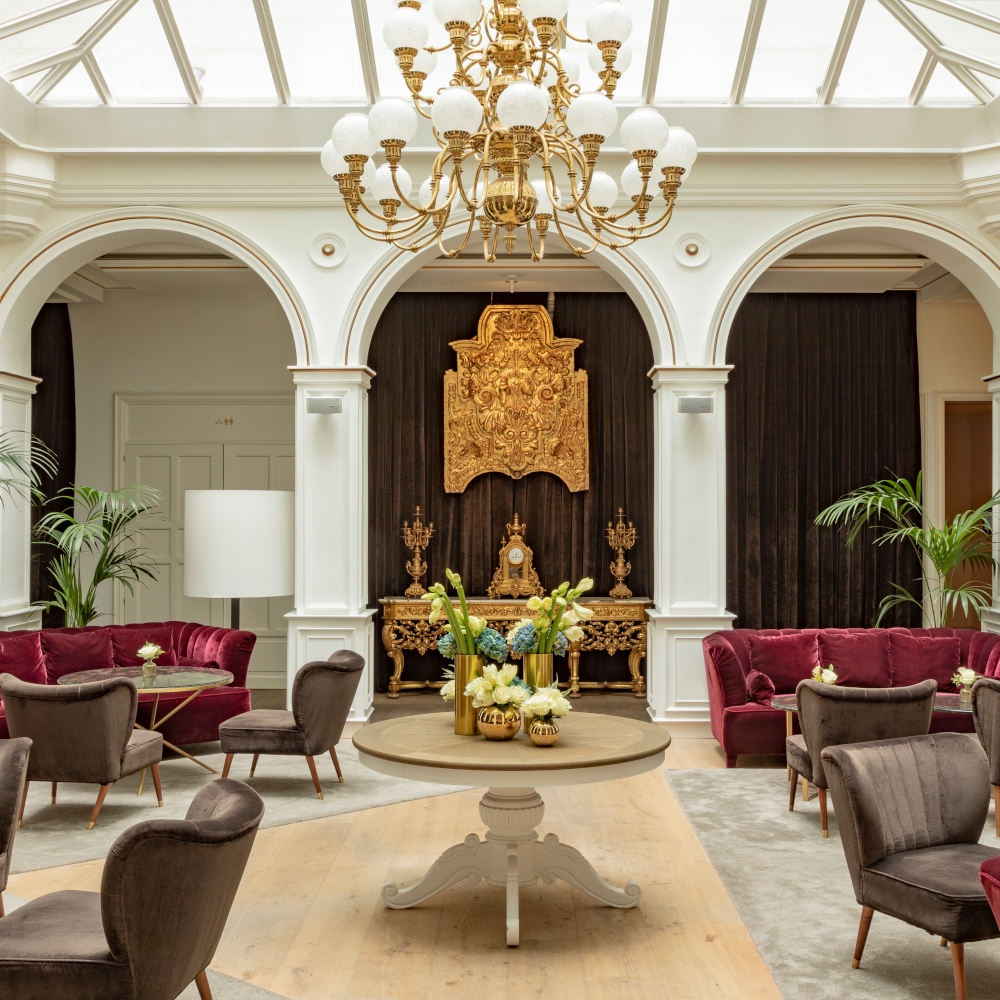 Bright interior of a lobby with roof windows, a large chandelier, arched doorways and red plush sofas.