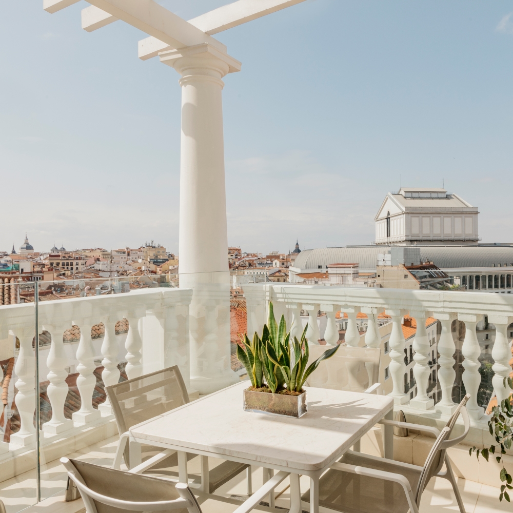 Exterior of a balcony with white pillar and railing and two chairs overlooking the rooftops and buildings of Madrid.