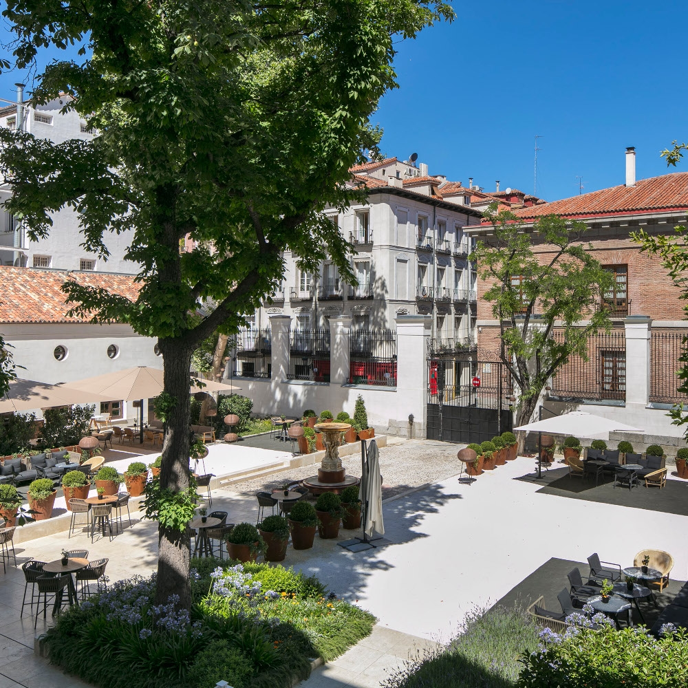 Open courtyard with trees, seats and dining chairs surrounded by traditional buildings with white and brick walls and red tile roofs.