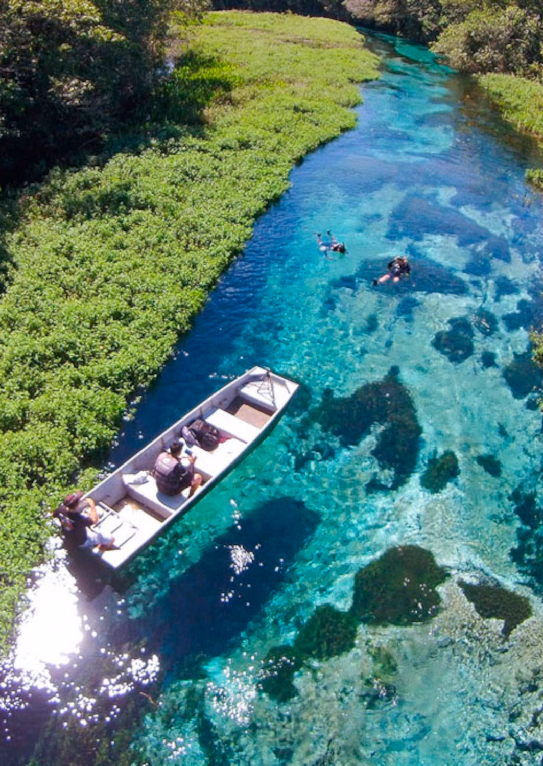 A clear blue river sidelined by greenery with a small boat and a few people swimming in the river.