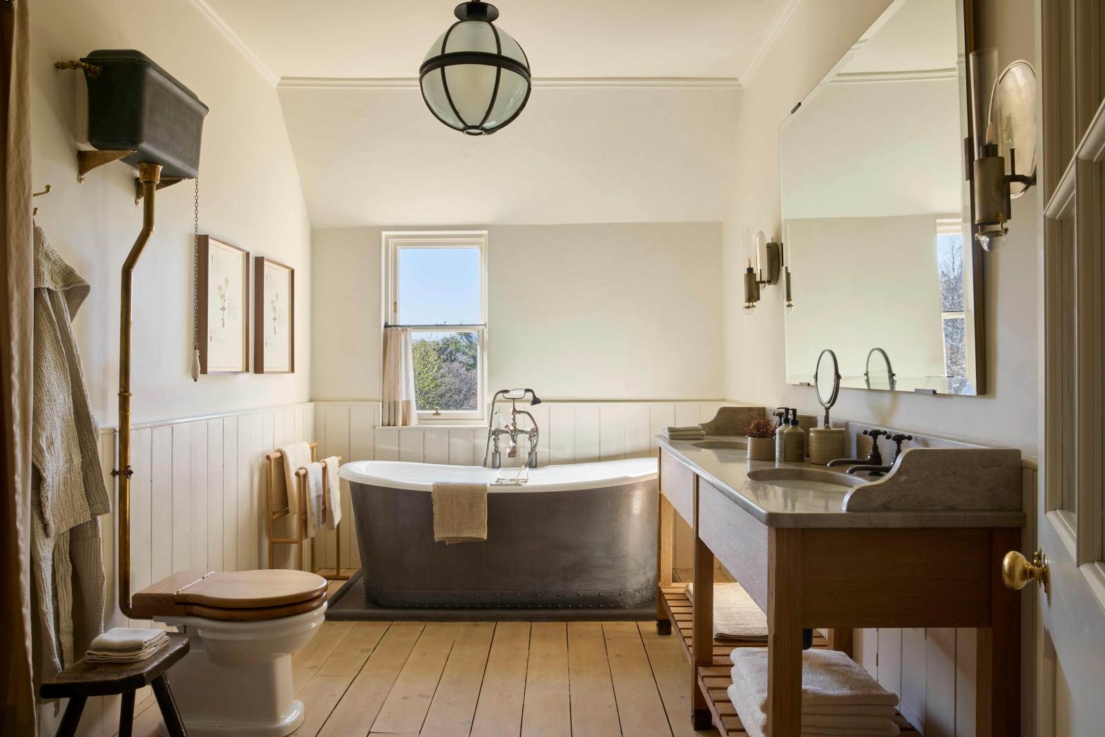 A large bathroom with a toilet, wooden floors, a double sink built into a cabinet and a large, freestanding bathtub by a small window.