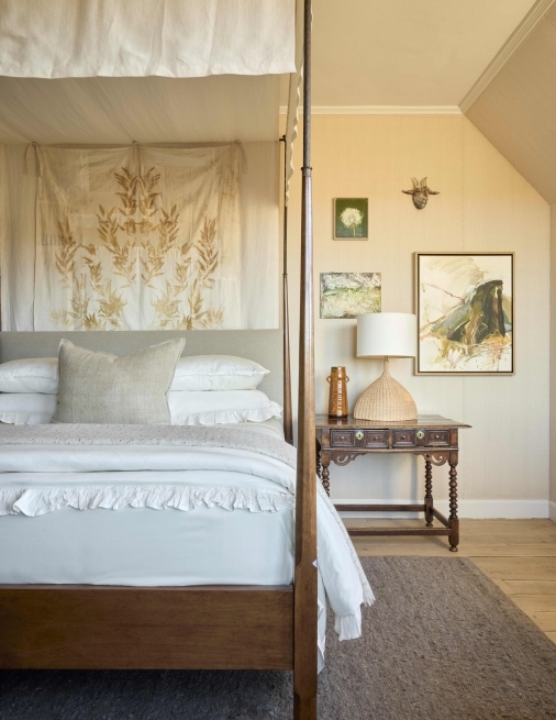 A traditional four-poster bed with white linens and a patterned sheet behind the headboard, with a bedside table, lamp and artworks on the wall.