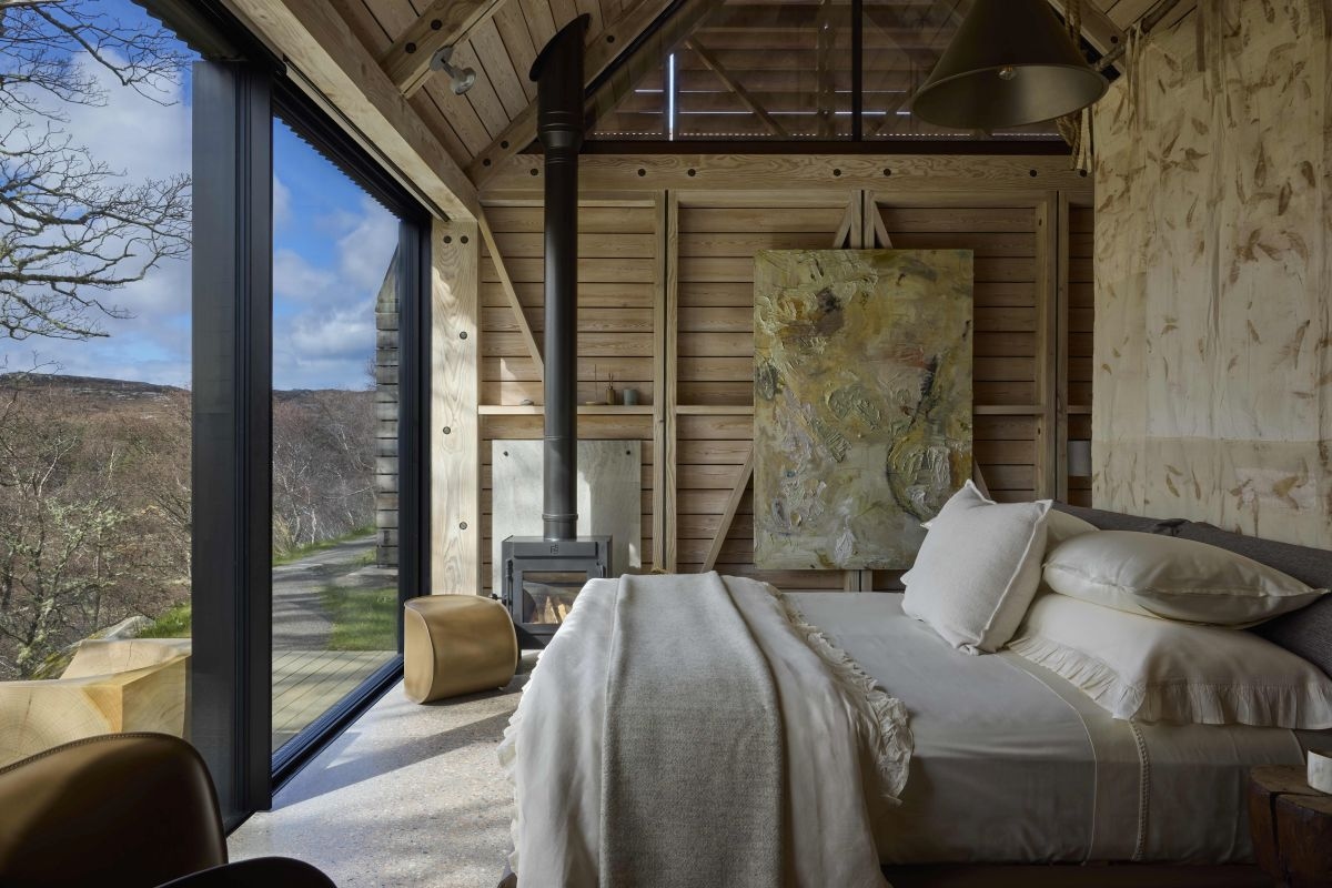 A luxurious king bed in a room with soft colours and wooden accents looks out through bi-fold glass doors to a wooden deck and trees.