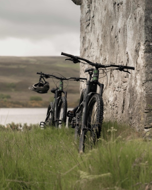 Two e-bikes, one in front of the other, leaning against a stone wall in the grass, with water views behind.