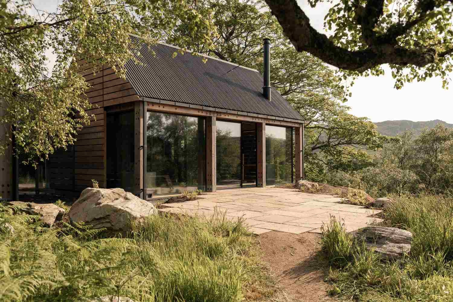 A timber, lodge-style cottage with black pitched roof, tall chimney and floor-to-ceiling glass at the front, leading to a paved terrace and surrounded by trees.