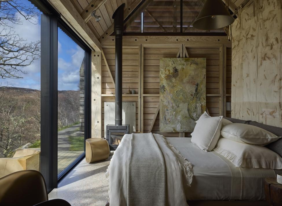 A luxurious king bed in a room with soft colours and wooden accents looks out through bi-fold glass doors to a wooden deck and trees.