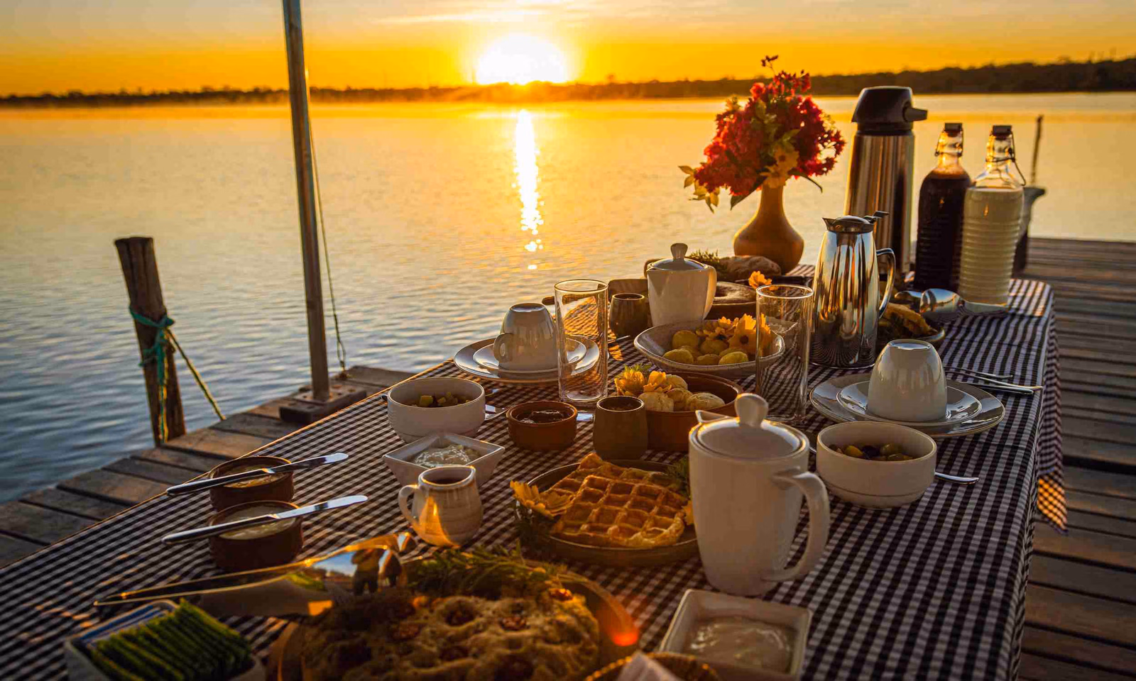 A table laid with a blue and white checked tablecloth and set with breakfast food and coffee, on a pier jutting over a lake as the sun rises in the background.